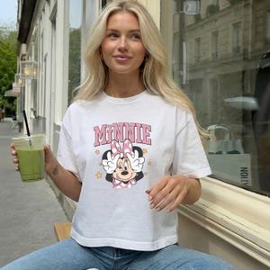 Pode incluir: Camiseta branca com a palavra "Minnie" em letras rosa e um desenho da Minnie Mouse. A Minnie Mouse dos desenhos animados tem um la&ccedil;o rosa e faz o sinal da paz. A camiseta tem mangas curtas.