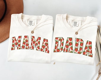 May include: Two off-white t-shirts with the words "MAMA" and "DADA" in a red strawberry pattern. Each shirt has a gold necklace with a round pendant. The shirts are folded and ready to wear.
