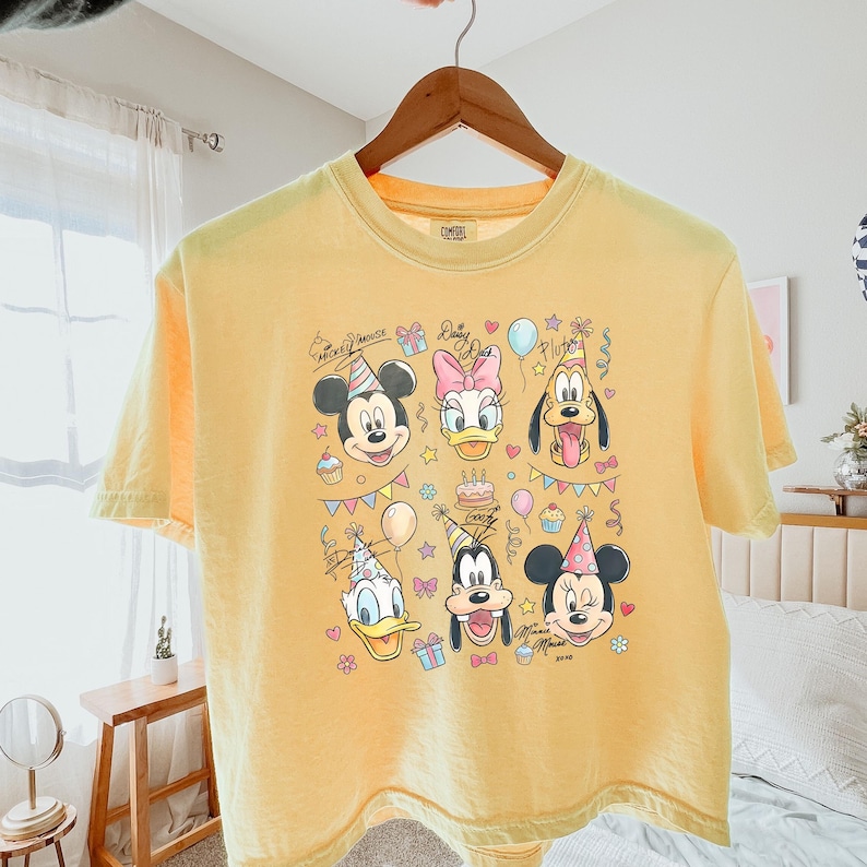 Op de afbeelding: Geel t-shirt met een kleurrijke afbeelding van Mickey Mouse, Daisy Duck, Pluto, Donald Duck, Goofy en Minnie Mouse, allemaal met feesthoeden. Het ontwerp bevat ballonnen, cupcakes en confetti. Het shirt hangt aan een houten hanger.