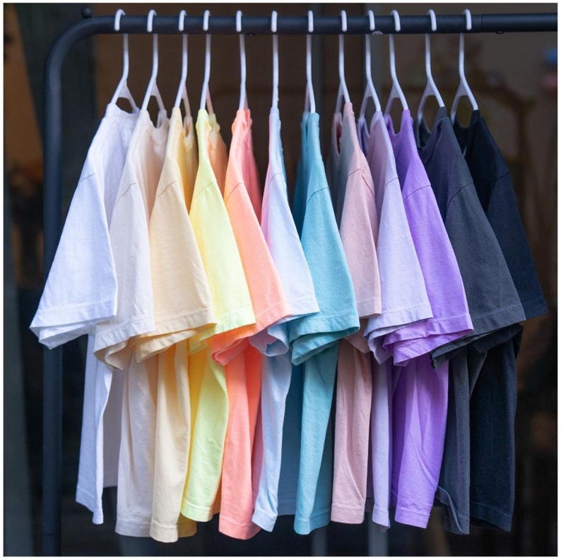Pode incluir: Uma cole&ccedil;&atilde;o de camisetas coloridas penduradas em um varal. As camisas est&atilde;o em v&aacute;rios tons pastel, incluindo branco, amarelo, p&ecirc;ssego, azul e roxo. As camisetas s&atilde;o feitas de um material macio e s&atilde;o exibidas em cabides brancos.