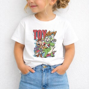 K&ouml;nnte beinhalten: Ein wei&szlig;es Kurzarm-T-Shirt mit einem farbenfrohen Grafikdruck der Toy Story-Figuren Woody, Buzz Lightyear und Rex. Das Design enth&auml;lt den Text Toy Story, Since 1995, Woody, Buzz und Rex in einer Schriftart im Retro-Stil.