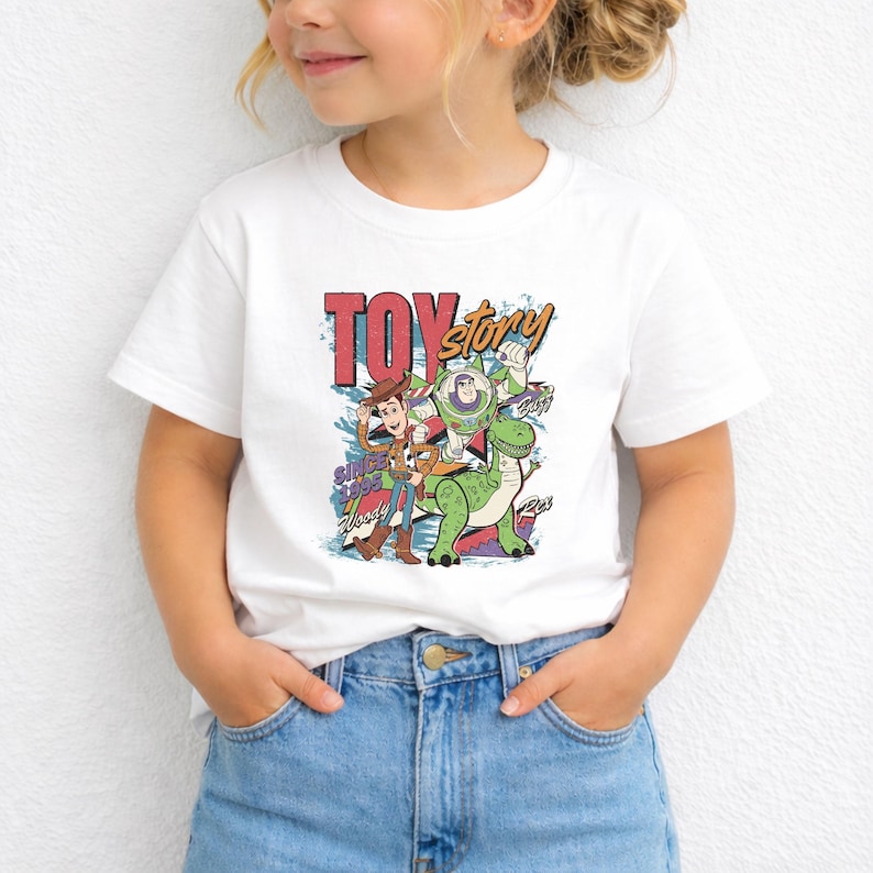 K&ouml;nnte beinhalten: Ein wei&szlig;es Kurzarm-T-Shirt mit einem farbenfrohen Grafikdruck der Toy Story-Figuren Woody, Buzz Lightyear und Rex. Das Design enth&auml;lt den Text Toy Story, Since 1995, Woody, Buzz und Rex in einer Schriftart im Retro-Stil.