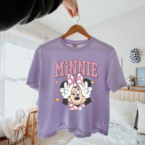 Pode incluir: Uma camiseta lil&aacute;s com um gr&aacute;fico da Minnie Mouse. A palavra "Minnie" &eacute; impressa em rosa acima da personagem. Minnie Mouse &eacute; retratada com um la&ccedil;o rosa de bolinhas e fazendo o sinal de paz. Estrelas amarelas circundam a personagem.