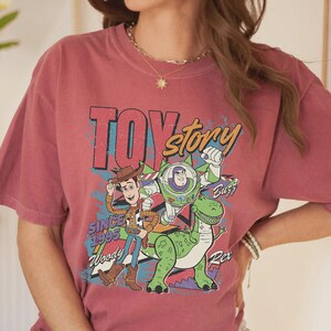 K&ouml;nnte beinhalten: Ein altrosa T-Shirt mit einem Grafikdruck der Toy Story-Figuren Woody, Buzz Lightyear und Rex. Das Design enth&auml;lt den Text "TOY story", "SINCE 1995", "Woody", "Buzz" und "Rex" in einem farbenfrohen, abgenutzten Retro-Stil.