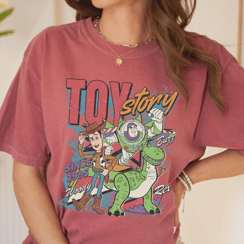 K&ouml;nnte beinhalten: Ein altrosa T-Shirt mit einem Grafikdruck der Toy Story-Figuren Woody, Buzz Lightyear und Rex. Das Design enth&auml;lt den Text "TOY story", "SINCE 1995", "Woody", "Buzz" und "Rex" in einem farbenfrohen, abgenutzten Retro-Stil.