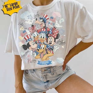 May include: White t-shirt featuring a colorful graphic of Disney characters Mickey Mouse, Minnie Mouse, Donald Duck, Daisy Duck, Goofy, and Pluto, celebrating the USA's 250th anniversary. The design includes American flags and fireworks.
