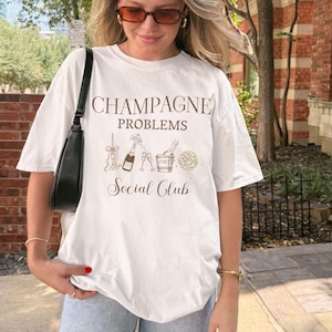 Può includere: T-shirt bianco con la scritta "CHAMPAGNE PROBLEMS Social Club" in marrone. La maglietta presenta una grafica di bottiglie di champagne, bicchieri e un secchiello. La maglietta è indossata da una persona.