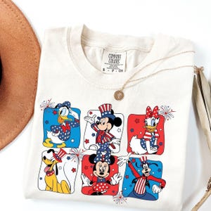May include: Cream-colored t-shirt featuring cartoon characters in patriotic outfits. Donald Duck, Mickey Mouse, Minnie Mouse, Daisy Duck, Pluto, and Goofy are depicted in red, white, and blue squares. A gold necklace and a brown hat are also visible.