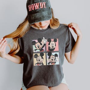 May include: Dark gray t-shirt featuring four panels with Mickey Mouse illustrations in various poses. The person is wearing a dark gray cap with the word "HOWDY" in red letters.