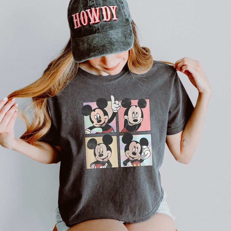 May include: Dark gray t-shirt featuring four panels with Mickey Mouse illustrations in various poses. The person is wearing a dark gray cap with the word "HOWDY" in red letters.