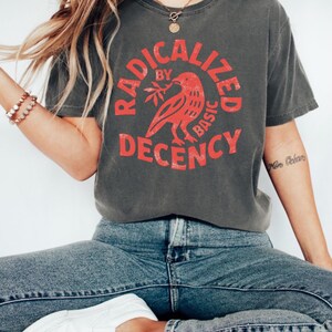 K&ouml;nnte beinhalten: Dunkelgraues T-Shirt mit dem Text "RADICALIZED BY BASIC DECENCY" in einem roten kreisf&ouml;rmigen Design. Das Design beinhaltet einen roten Vogel, der einen Zweig h&auml;lt. Das Shirt wird mit blauen Jeans und wei&szlig;en Schuhen getragen.