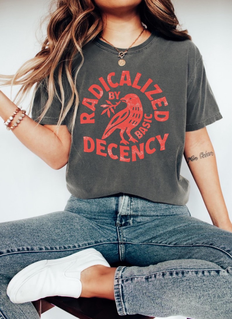 K&ouml;nnte beinhalten: Dunkelgraues T-Shirt mit dem Text "RADICALIZED BY BASIC DECENCY" in einem roten kreisf&ouml;rmigen Design. Das Design beinhaltet einen roten Vogel, der einen Zweig h&auml;lt. Das Shirt wird mit blauen Jeans und wei&szlig;en Schuhen getragen.