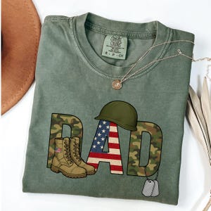 Könnte beinhalten: Olivgrünes Comfort Colors T-Shirt mit einem "DAD"-Motiv im Camouflage-Design. Das "A" zeigt die amerikanische Flagge, mit Militärhelm, Stiefeln und Erkennungsmarken. Eine silberne Halskette liegt über dem Shirt.