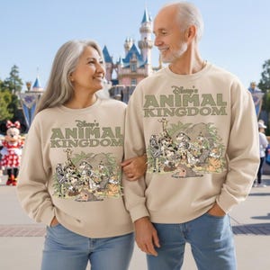 May include: Beige sweatshirts with the text "Disney's Animal Kingdom" and a graphic of various cartoon animals. The sweatshirts are worn by a couple in front of a castle and a Minnie Mouse character.