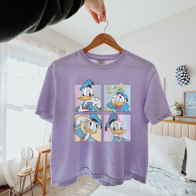 May include: A light purple t-shirt featuring four panels with different expressions of Donald Duck. The cartoon character is wearing a blue sailor suit and hat. The shirt is hanging on a wooden hanger.