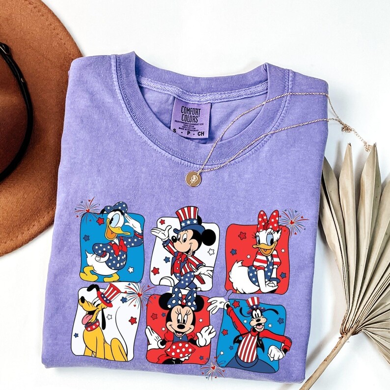 May include: A lavender Comfort Colors t-shirt featuring a patriotic design with cartoon characters. The design includes Mickey Mouse, Minnie Mouse, Donald Duck, Daisy Duck, Pluto, and Goofy in red, white, and blue outfits. A gold necklace is draped across the shirt.