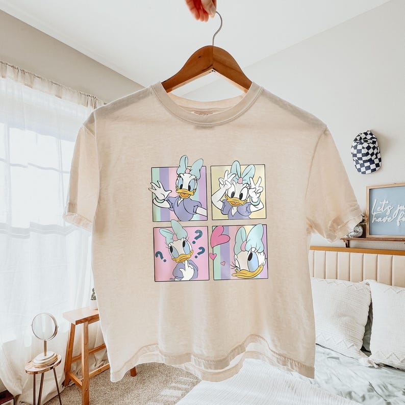 May include: Cream-colored t-shirt featuring a four-panel graphic of Daisy Duck in various poses. Each panel has a different pastel background. The shirt is hanging on a wooden hanger.