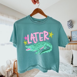 Könnte beinhalten: Türkisfarbenes T-Shirt mit dem Wort "LATER" in Rosa, weiß umrandet, und gelben Sternen. Darunter eine grüne Alligator-Illustration mit rosa Zähnen. Das T-Shirt hängt an einem Holzbügel.