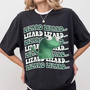May include: Black t-shirt featuring a green cartoon lizard with large eyes and the text "LIZARD LIZARD..." repeated in white and green. The lizard is waving. The shirt has a crew neck and short sleeves.