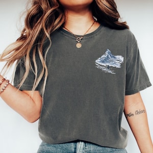 Puede incluir: Camiseta gris oscuro con un gr&aacute;fico de monta&ntilde;a en tonos azules y blancos en la parte delantera. La camiseta tiene mangas cortas y cuello redondo. La tela parece suave y c&oacute;moda, adecuada para uso casual.