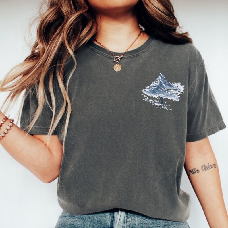 Puede incluir: Camiseta gris oscuro con un gr&aacute;fico de monta&ntilde;a en tonos azules y blancos en la parte delantera. La camiseta tiene mangas cortas y cuello redondo. La tela parece suave y c&oacute;moda, adecuada para uso casual.