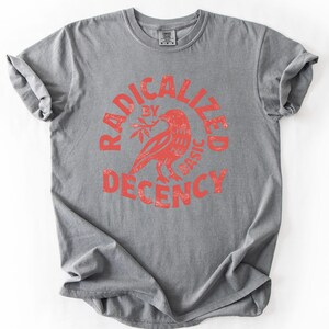 K&ouml;nnte beinhalten: Graues T-Shirt mit roter Grafik. Die Grafik zeigt einen Vogel, der einen Zweig in einem Kreis h&auml;lt, mit den Worten "RADICALIZED BY BASIC DECENCY" in einer verwitterten Schriftart. Das T-Shirt hat kurze &Auml;rmel und einen Rundhalsausschnitt.