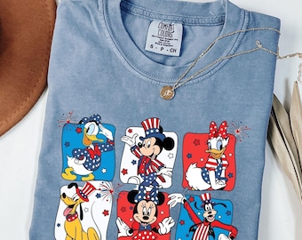 May include: A light blue Comfort Colors t-shirt featuring a patriotic design with Disney characters. The graphic includes Donald Duck, Mickey Mouse, Daisy Duck, Pluto, Minnie Mouse, and Goofy, each in red, white, and blue attire. A gold necklace rests on the shirt.