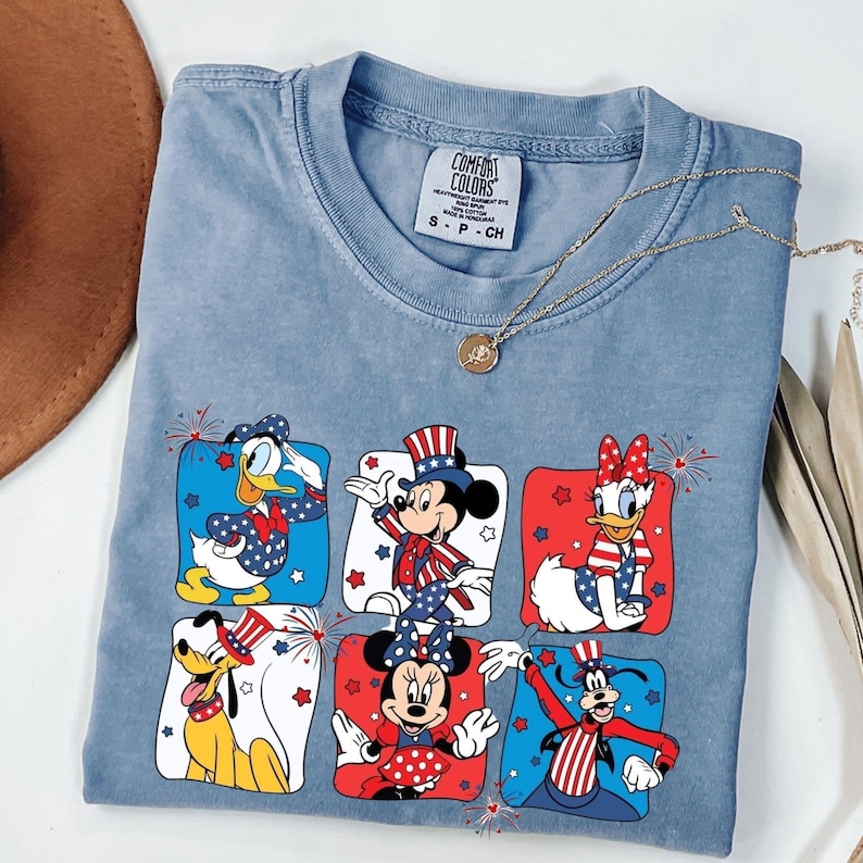 May include: A light blue Comfort Colors t-shirt featuring a patriotic design with Disney characters. The graphic includes Donald Duck, Mickey Mouse, Daisy Duck, Pluto, Minnie Mouse, and Goofy, each in red, white, and blue attire. A gold necklace rests on the shirt.
