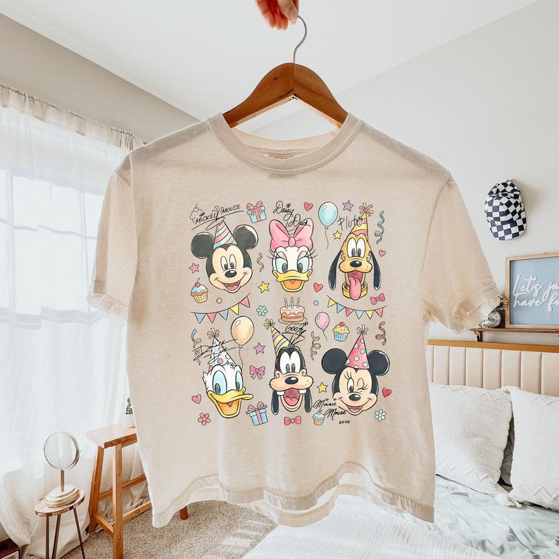 Op de afbeelding: Een beige t-shirt met een kleurrijke afbeelding van Mickey Mouse, Daisy Duck, Pluto, Donald Duck, Goofy en Minnie Mouse, elk met een feesthoed. Het ontwerp bevat cupcakes, ballonnen en een taart, met de woorden "Mickey Mouse" en "Daisy" in script.