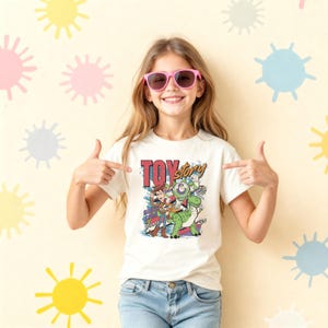K&ouml;nnte beinhalten: Ein wei&szlig;es kurz&auml;rmeliges T-Shirt mit einem farbenfrohen Grafikdruck der Toy Story-Figuren Woody, Buzz Lightyear und Rex. Der Text Toy Story ist in Rot und Orange &uuml;ber den Figuren gedruckt, erg&auml;nzt durch blaue und gr&uuml;ne Akzente.