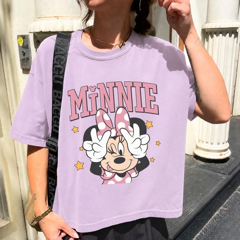 Pode incluir: Camiseta lil&aacute;s claro com um gr&aacute;fico da Minnie Mouse. O design inclui a palavra "MINNIE" em rosa, Minnie Mouse com um la&ccedil;o rosa e estrelas amarelas. Minnie faz um sinal de paz com as m&atilde;os.