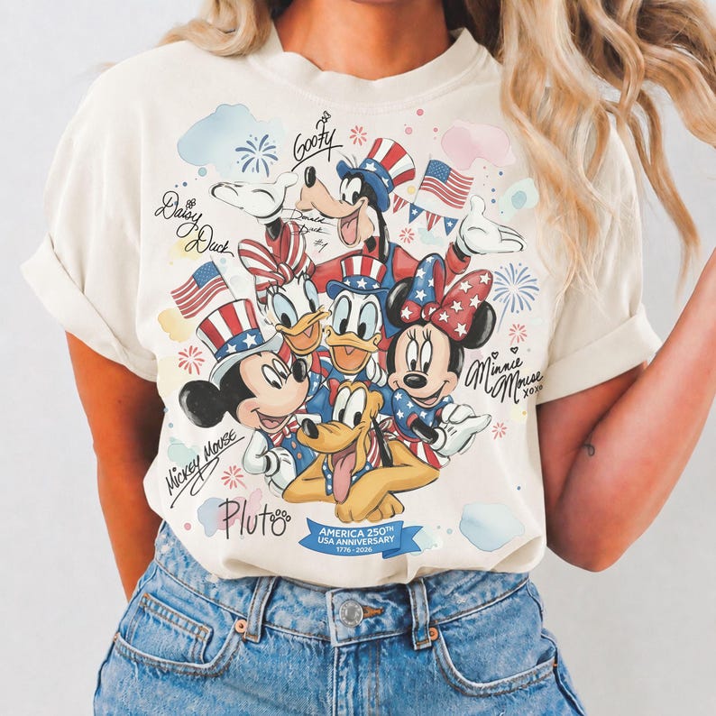 May include: Cream-colored t-shirt featuring a graphic of Disney characters Mickey Mouse, Minnie Mouse, Donald Duck, Daisy Duck, Goofy, and Pluto. The design includes American flags, fireworks, and text that reads "America 250th USA Anniversary 1776-2026."