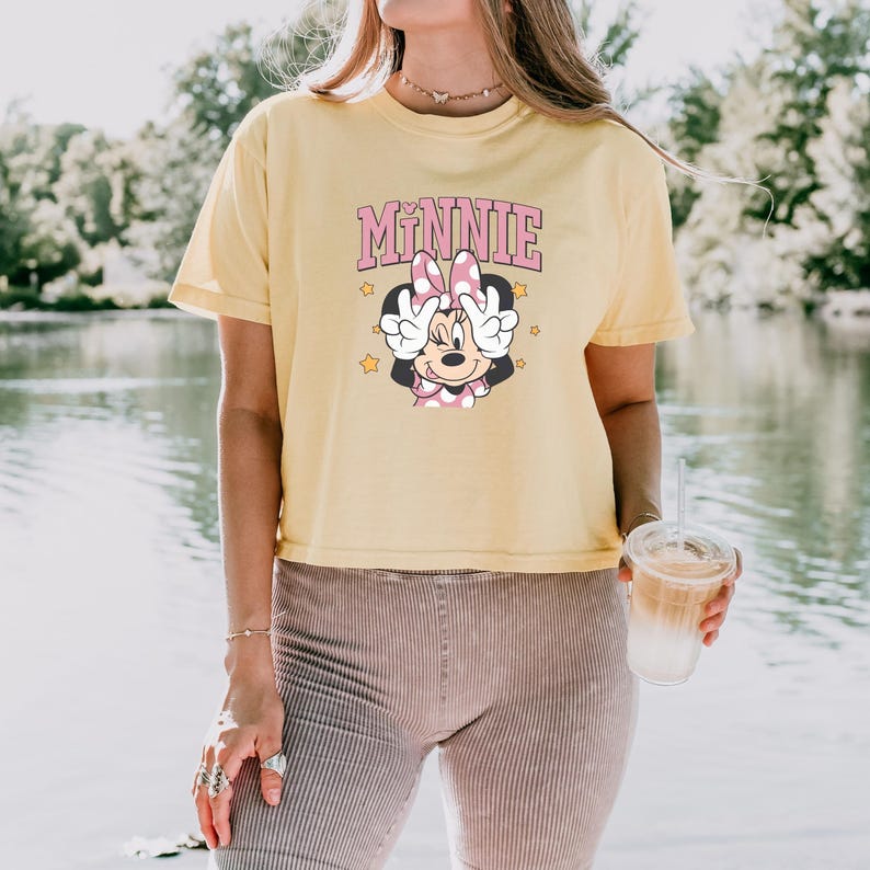 Pode incluir: Camiseta amarelo claro com um desenho da Minnie Mouse. O desenho inclui o nome "Minnie" em letras rosa acima de uma imagem de desenho animado da Minnie Mouse fazendo o sinal de paz. A camiseta &eacute; de manga curta.