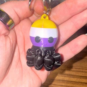 May include: A keychain featuring a small octopus with a non-binary pride flag design. The octopus has a yellow top, white middle, and purple bottom, with black tentacles. The keychain is attached to a silver keyring.