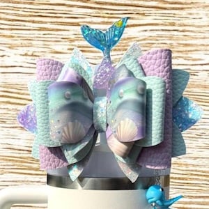 May include: A mermaid-themed hair bow in shades of blue, purple, and teal, with a glittery finish. The bow features a seashell design and a blue mermaid tail accent. A small blue whale charm hangs below.