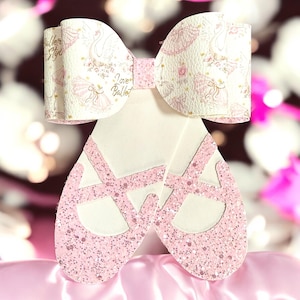 May include: A decorative bow and ballet slipper set. The bow is white with a pink ballet-themed pattern and the words "Baller Style" and "I love Ballet". The ballet slippers are white with pink glitter accents.