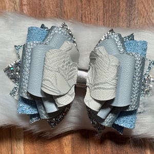 May include: A layered hair bow in shades of light blue, silver, and white. The bow features textured and glittery materials, with a silver center. The bow is set against a white, fluffy background.
