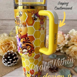 Puede incluir: Vaso t&eacute;rmico aislado amarillo y dorado con personajes de Winnie the Pooh y Tigger, con un patr&oacute;n de panal. El vaso tiene un asa amarilla con el nombre "Jackie" y el texto "Saying Customised" y "Craft Within design 2".