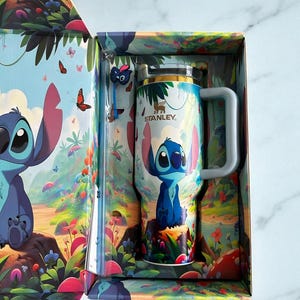 Puede incluir: Una taza Stanley azul claro con un dise&ntilde;o de personaje de dibujos animados, presentada en una caja a juego. La taza tiene un asa y una tapa plateada. La caja y la taza presentan un dise&ntilde;o colorido con el texto "Light Blue Cup" y "CraftWithin".