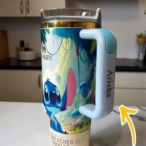 Puede incluir: Vaso t&eacute;rmico con dise&ntilde;o de Stitch. El vaso tiene un asa azul claro con el nombre "Arisha" impreso. El vaso est&aacute; etiquetado como "Quencher H2.0 Tumbler". Ideal para bebidas fr&iacute;as.