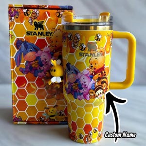 Puede incluir: Vaso Stanley amarillo con asa, con un patr&oacute;n de panal y im&aacute;genes de Winnie the Pooh y sus amigos. El vaso tiene una tapa y un asa amarillas, y el texto "STANLEY" es visible. La caja de al lado tiene un dise&ntilde;o a juego. El texto "CraftWithin" est&aacute; en la parte superior.