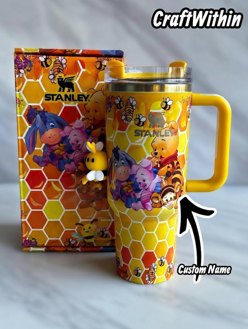 Puede incluir: Vaso Stanley amarillo con asa, con un patr&oacute;n de panal y im&aacute;genes de Winnie the Pooh y sus amigos. El vaso tiene una tapa y un asa amarillas, y el texto "STANLEY" es visible. La caja de al lado tiene un dise&ntilde;o a juego. El texto "CraftWithin" est&aacute; en la parte superior.
