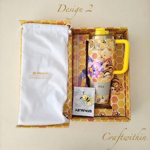 Puede incluir: Un vaso Stanley amarillo y beige con un dise&ntilde;o de Winnie the Pooh, un asa amarilla y una pajita a juego, presentado en una caja decorativa. La caja presenta un patr&oacute;n de panal y incluye una bolsa blanca con cord&oacute;n.