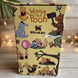Puede incluir: Una caja Stanley amarilla con personajes de Winnie the Pooh. La caja presenta ilustraciones de Pooh, Eeyore, Tigger y Piglet. El texto en la caja incluye "Winnie the Pooh", "Bounce This Way" y "Happy Birthday".