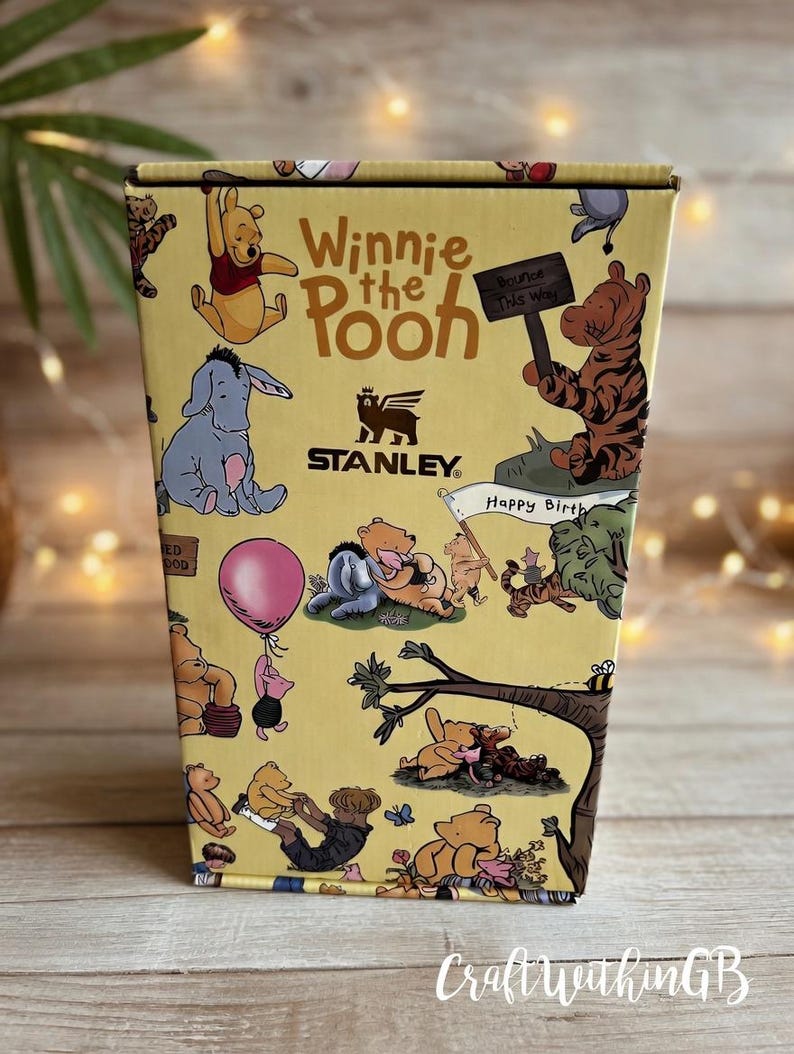 Puede incluir: Una caja Stanley amarilla con personajes de Winnie the Pooh. La caja presenta ilustraciones de Pooh, Eeyore, Tigger y Piglet. El texto en la caja incluye "Winnie the Pooh", "Bounce This Way" y "Happy Birthday".