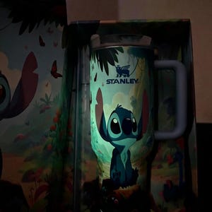 Puede incluir: Un termo de viaje Stanley con un personaje de dibujos animados, Stitch, en una escena de la jungla. El termo tiene un asa azul y el texto "STANLEY". Las palabras "Glow In The Dark" son visibles. El termo est&aacute; dentro de una caja con un dise&ntilde;o a juego.