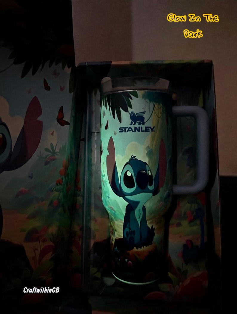 Puede incluir: Un termo de viaje Stanley con un personaje de dibujos animados, Stitch, en una escena de la jungla. El termo tiene un asa azul y el texto "STANLEY". Las palabras "Glow In The Dark" son visibles. El termo est&aacute; dentro de una caja con un dise&ntilde;o a juego.