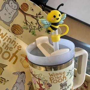 Puede incluir: Una taza tem&aacute;tica de Winnie the Pooh con un adorno de pajita de abeja amarilla y negra. La taza tiene un asa blanca y una tapa. La taza presenta ilustraciones de Winnie the Pooh y sus amigos. El texto "Winnie the Pooh" es visible.