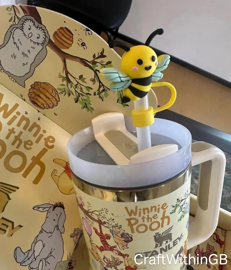 Puede incluir: Una taza tem&aacute;tica de Winnie the Pooh con un adorno de pajita de abeja amarilla y negra. La taza tiene un asa blanca y una tapa. La taza presenta ilustraciones de Winnie the Pooh y sus amigos. El texto "Winnie the Pooh" es visible.
