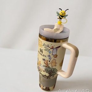 Puede incluir: Un termo de viaje Stanley beige con personajes de Winnie the Pooh, con borde y asa dorados. El termo tiene una tapa transparente y un adorno de pajita con forma de abeja amarilla. El termo est&aacute; decorado con ilustraciones de Winnie the Pooh y sus amigos.